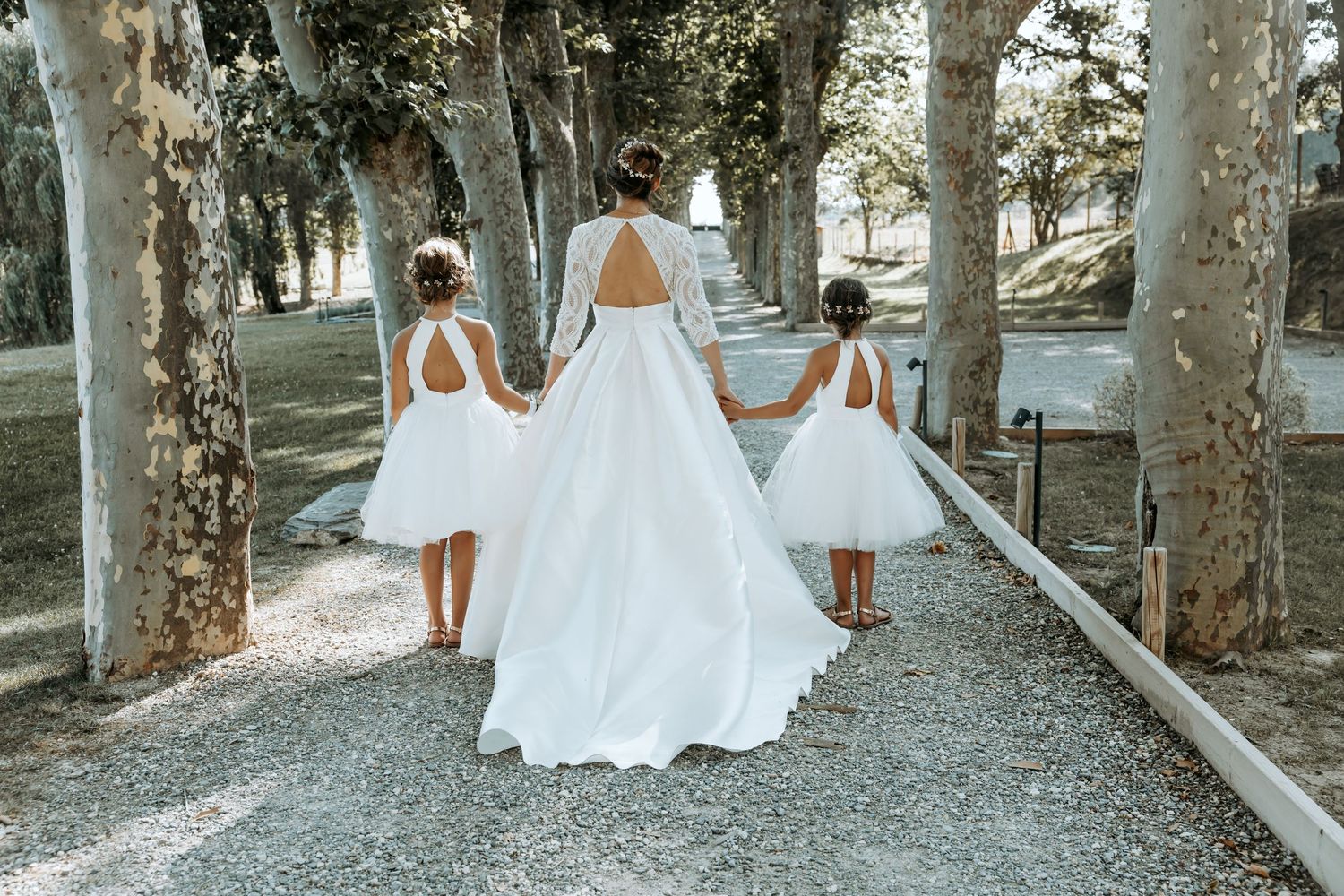 Bridal party in white dresses walking through tree-lined garden path Domaine la Plume photographe mariage