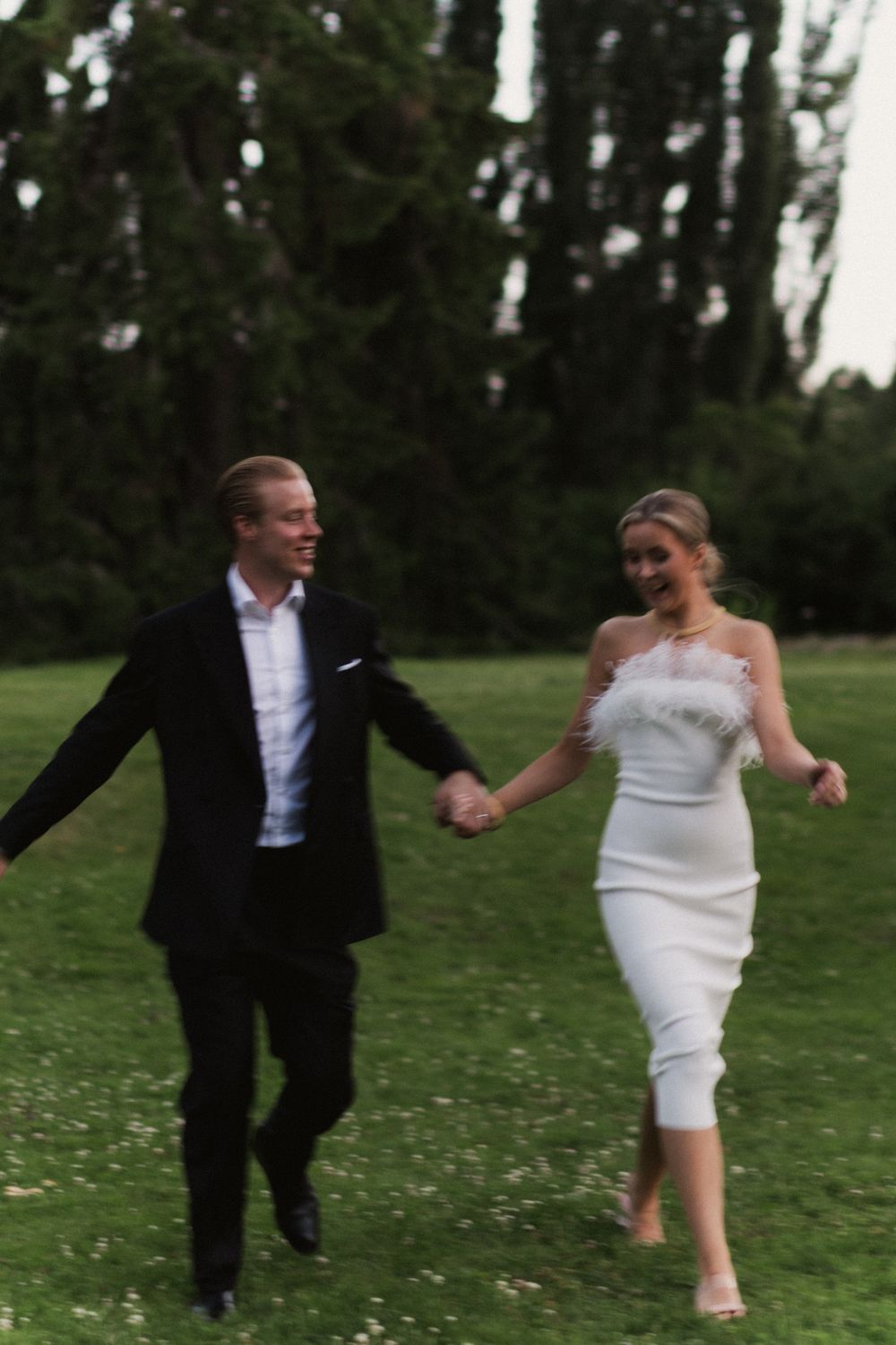 Playful couple running through a grassy field in wedding attire.