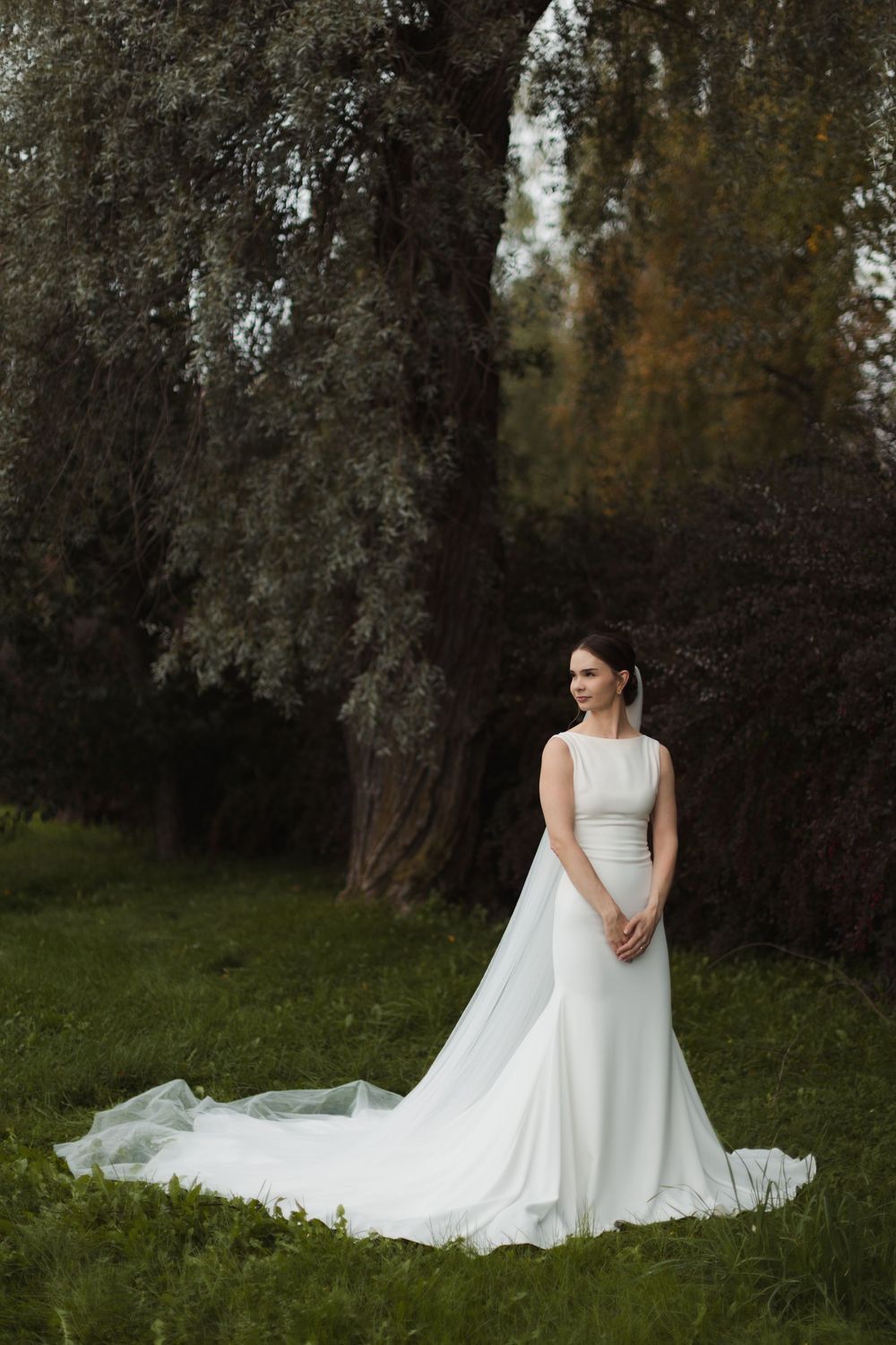 Elegant bride in flowing white gown poses in garden setting with autumn trees.