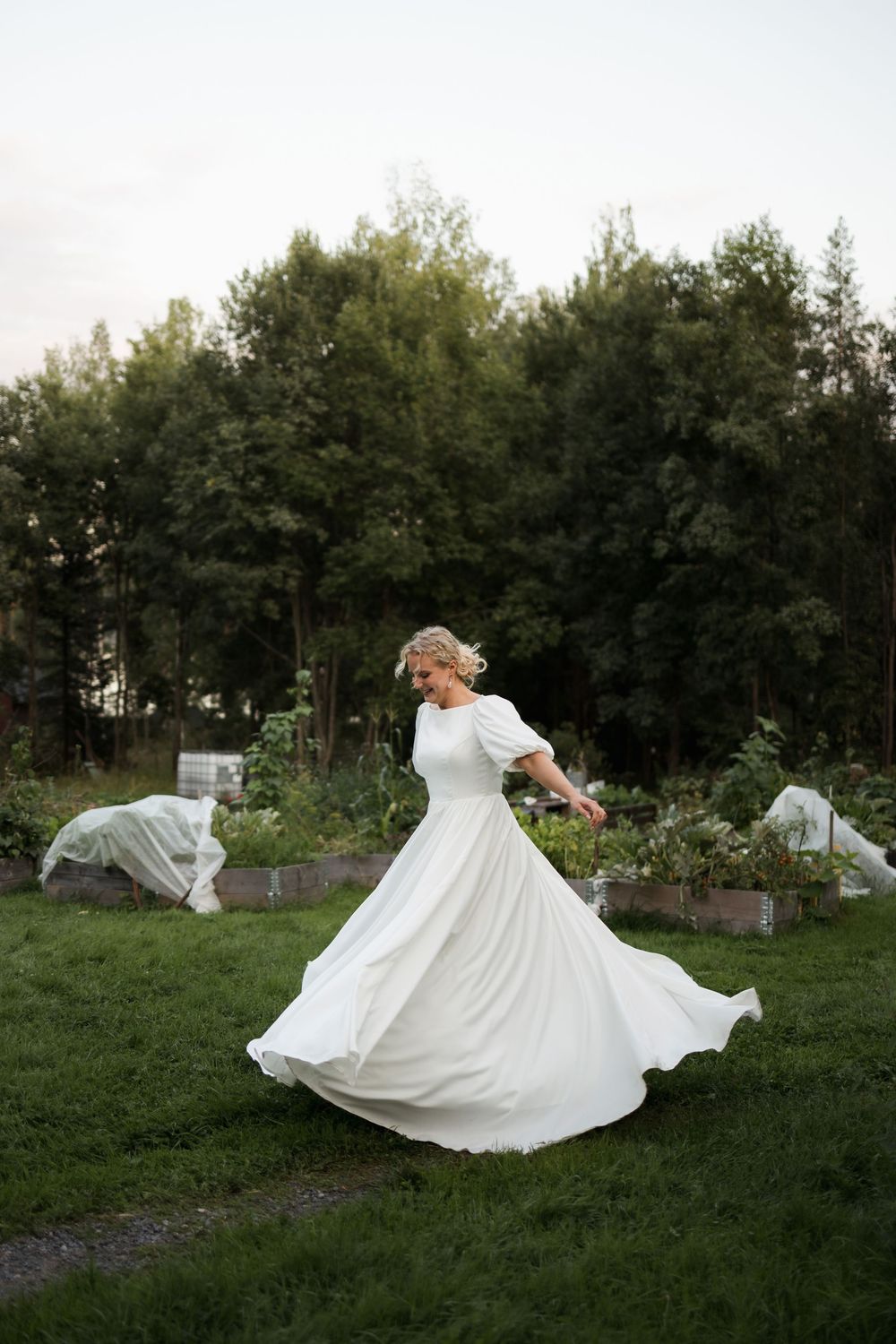 Bride in flowing white dress twirling in garden with trees in background.