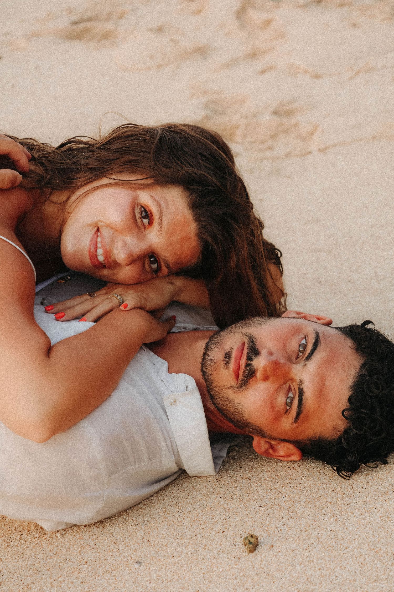 Couple lying on sandy beach laughing, woman resting on man's back in a playful and joyful pose.
