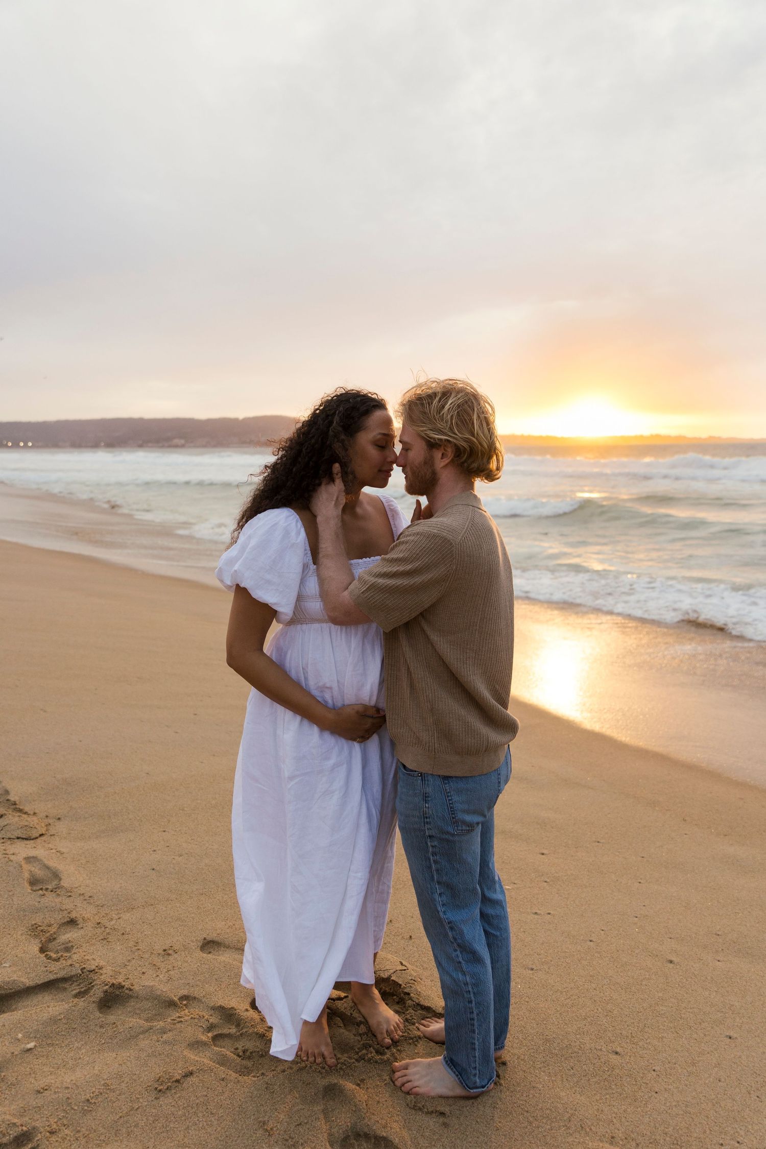 A couple shares an intimate moment on a sandy beach at sunset with waves crashing behind them.