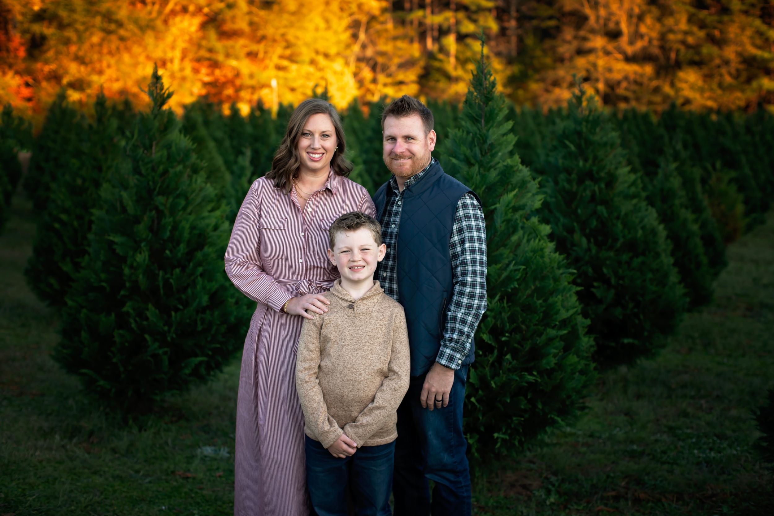 A family poses together at a Christmas tree farm with vibrant autumn foliage in the background.