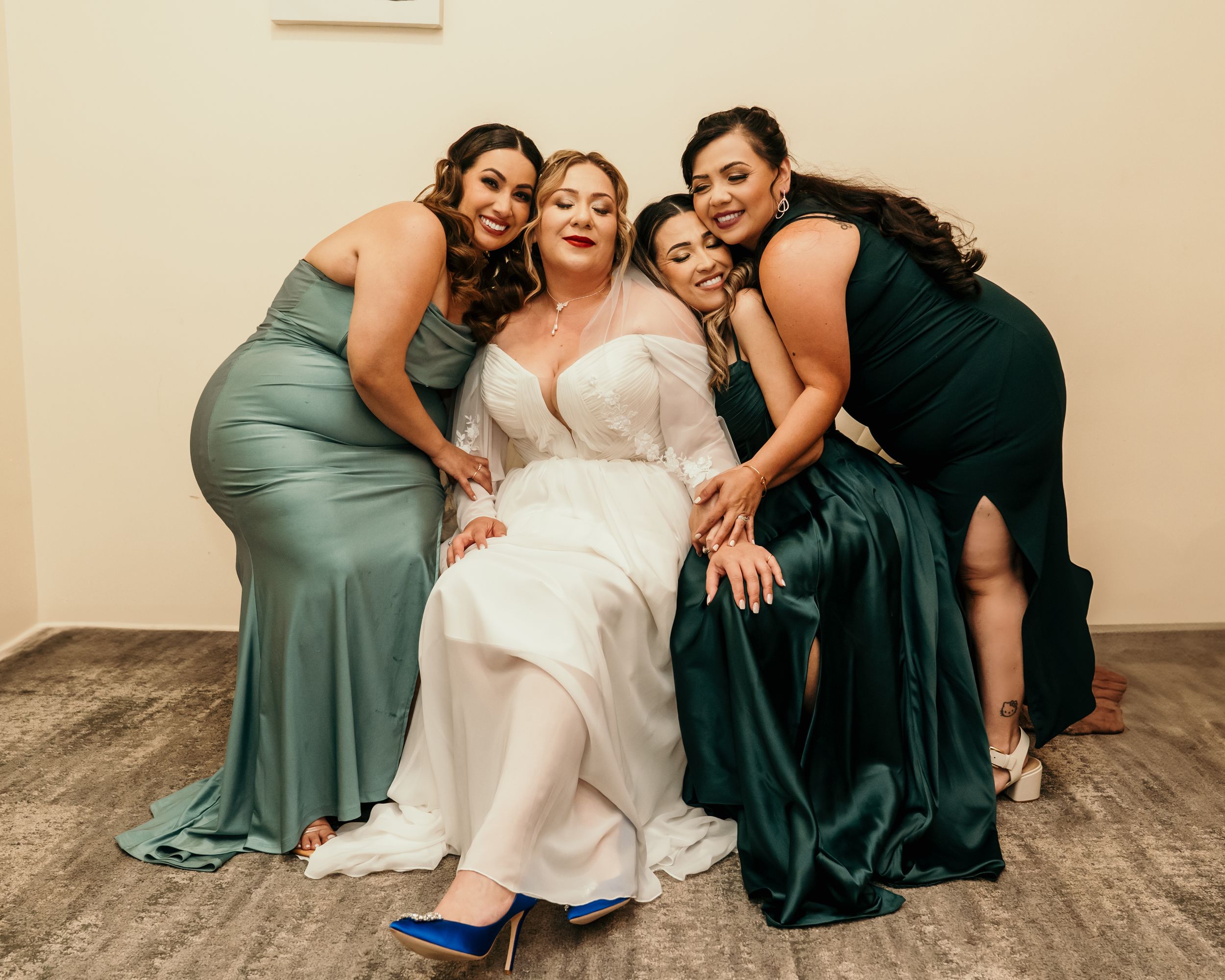 Bridesmaids in green and black dresses help adjust the bride's white gown while laughing together.
