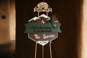 A rustic wooden welcome sign for 'The Gardens' features a tree silhouette and mountain design in dark green and brown tones.