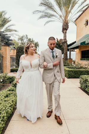 A couple walking together outdoors with palm trees, wearing formal wedding attire.