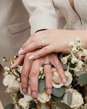 Close-up of wedding rings and hands resting on white roses and eucalyptus arrangement.
