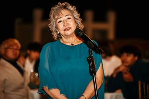 A woman in a teal dress speaks into a microphone during an evening event.