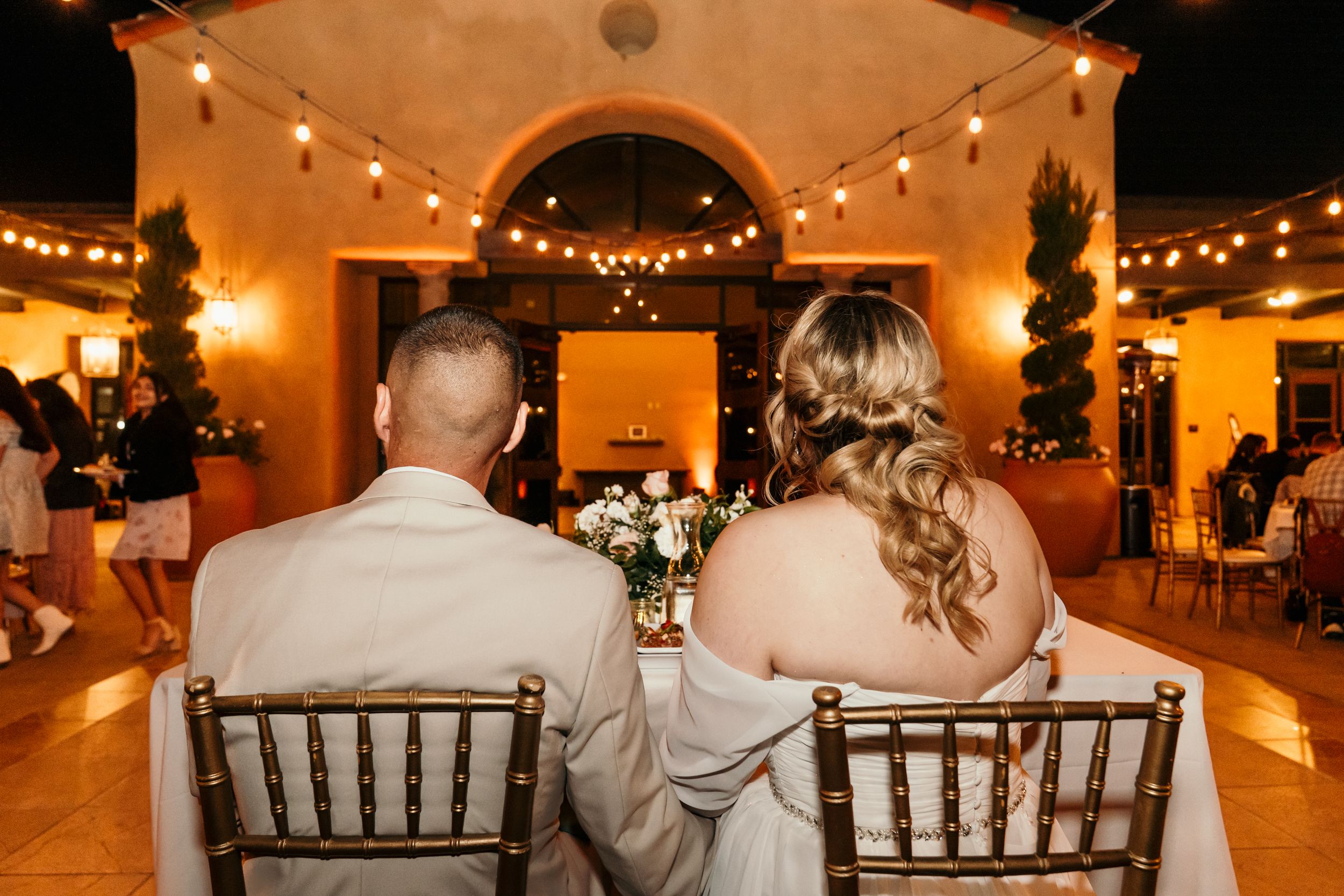 A couple sits together at their wedding reception under warm string lights.