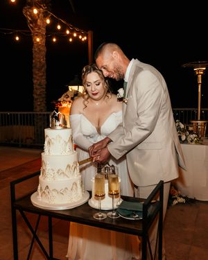 A couple cuts their elegant white and gold wedding cake together under string lights.