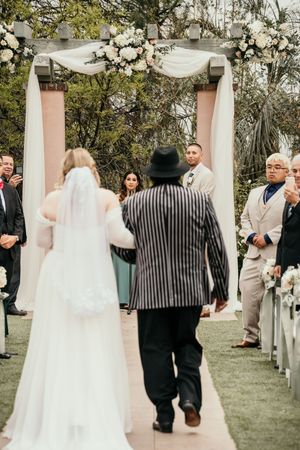 A wedding ceremony takes place under an elegant white-draped arch decorated with white floral arrangements.