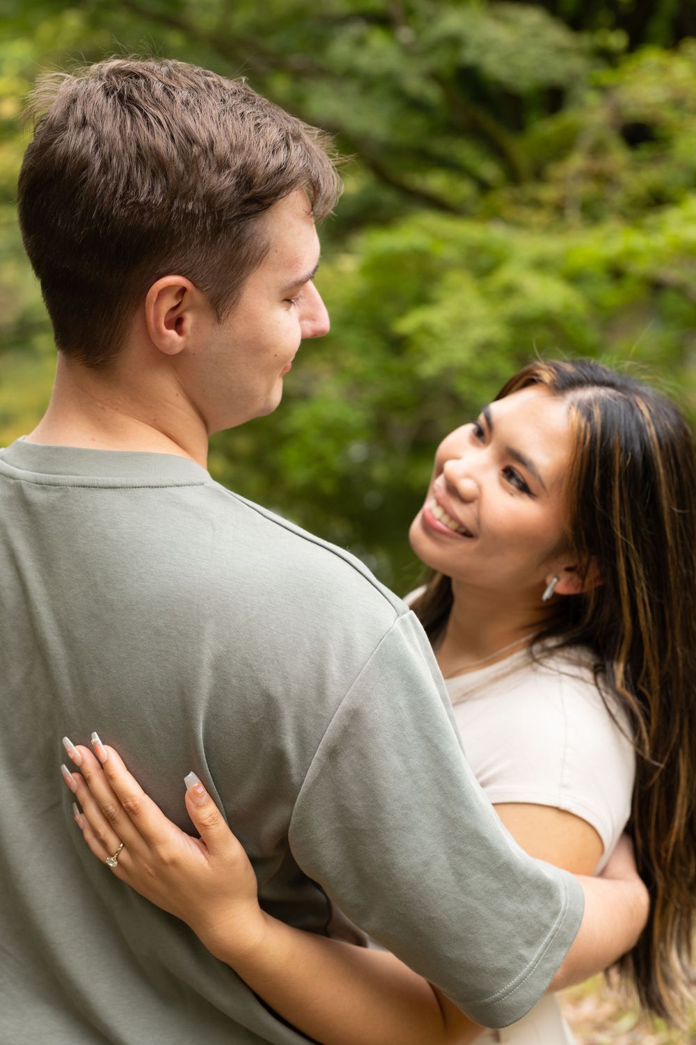 Woman smiling at her partner outdoors, showing engagement ring with green trees behind them.