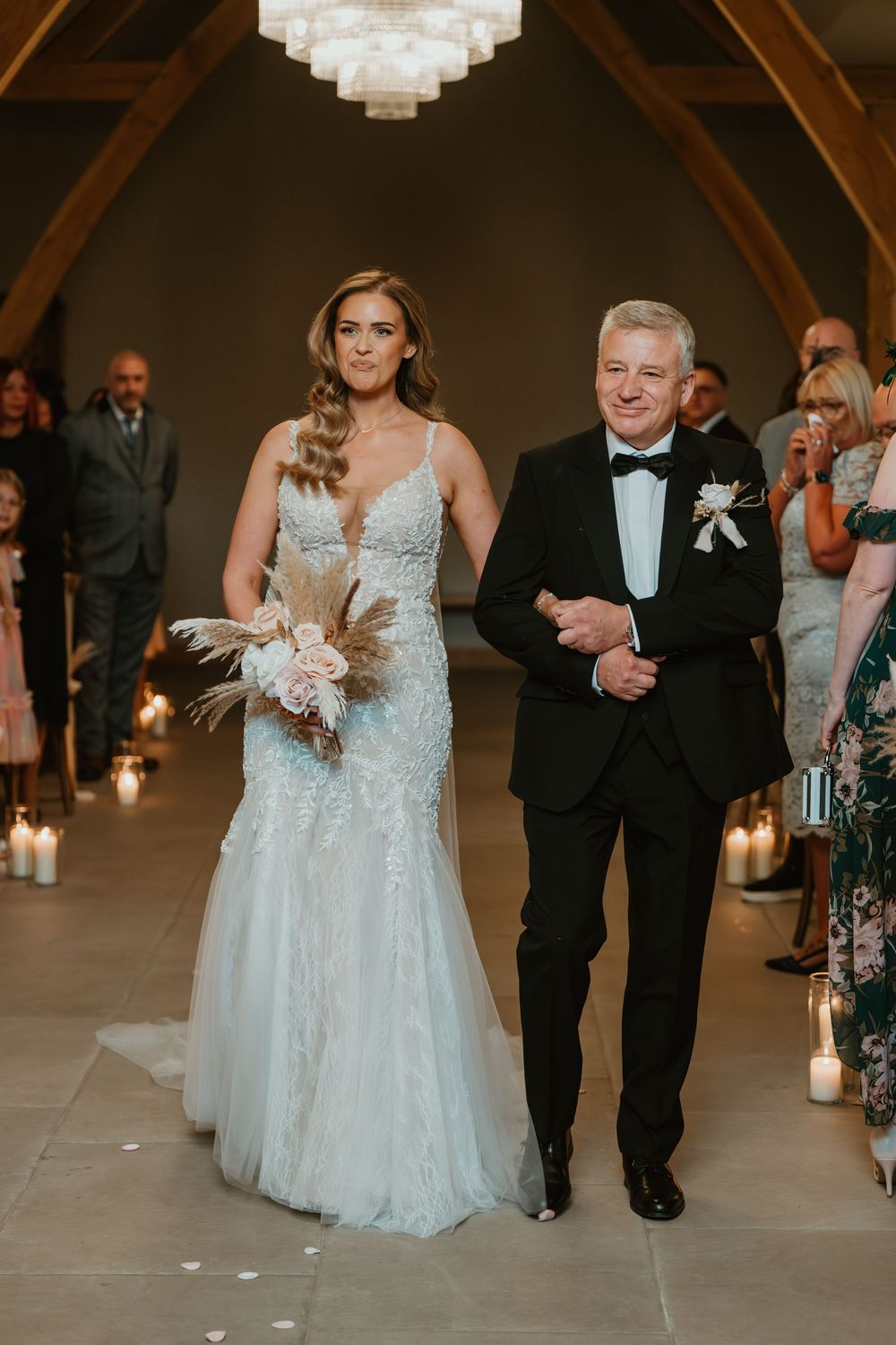 A bride in a white lace gown walks down the aisle with an escort in a black suit during an indoor wedding ceremony.