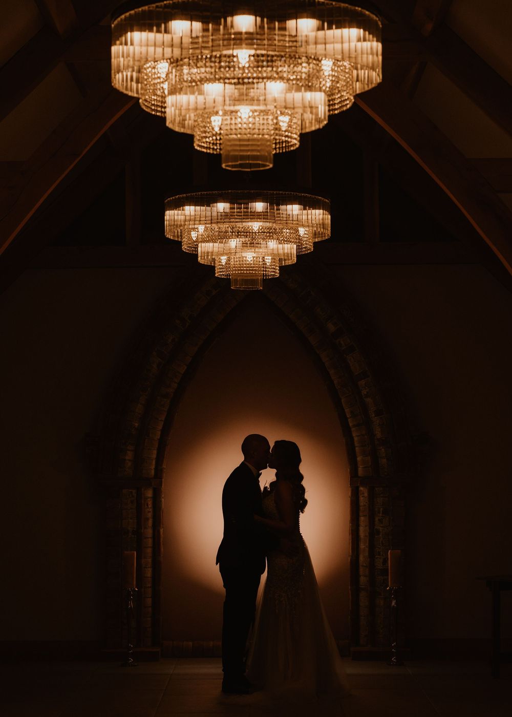 Silhouetted couple stands beneath elegant crystal chandeliers in a dark arched hallway creating a romantic ambiance.