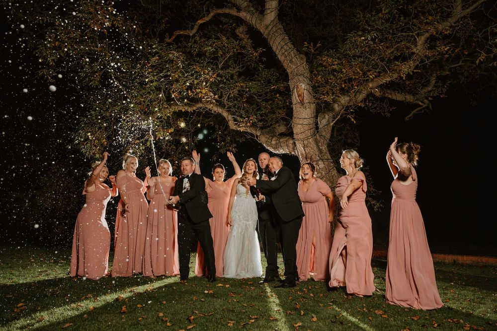 Wedding party members celebrate with raised arms under twinkling lights in an outdoor evening setting.