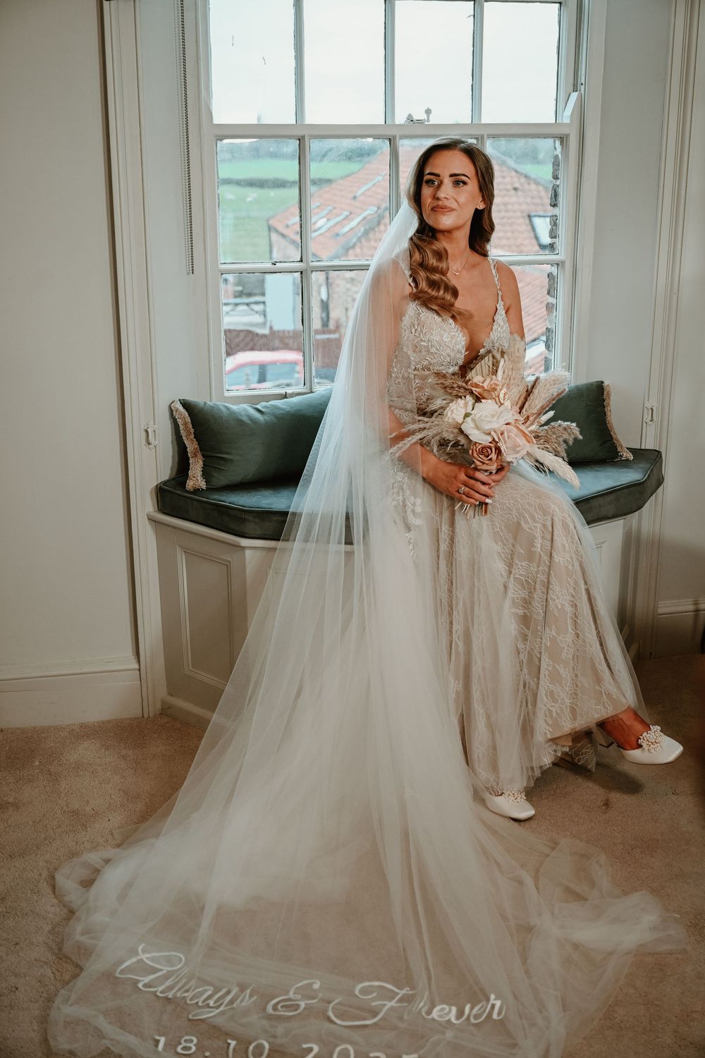 A bride in a flowing white wedding gown sits elegantly by a window with natural light streaming in.