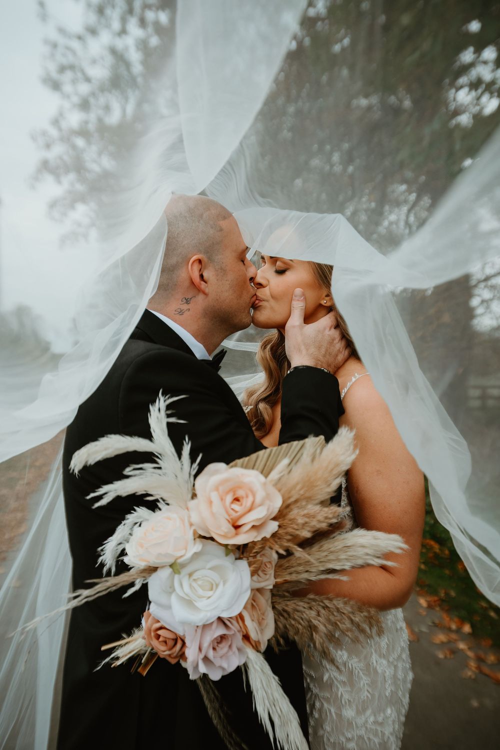 Romantic wedding couple embraces with flowing veil and blush pink floral bouquet with pampas grass in dreamy setting.