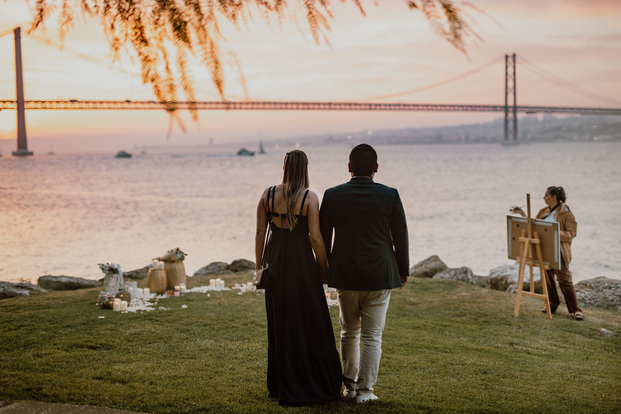 Silhouettes of people standing on a grassy hill at sunset with a bridge visible in the background.