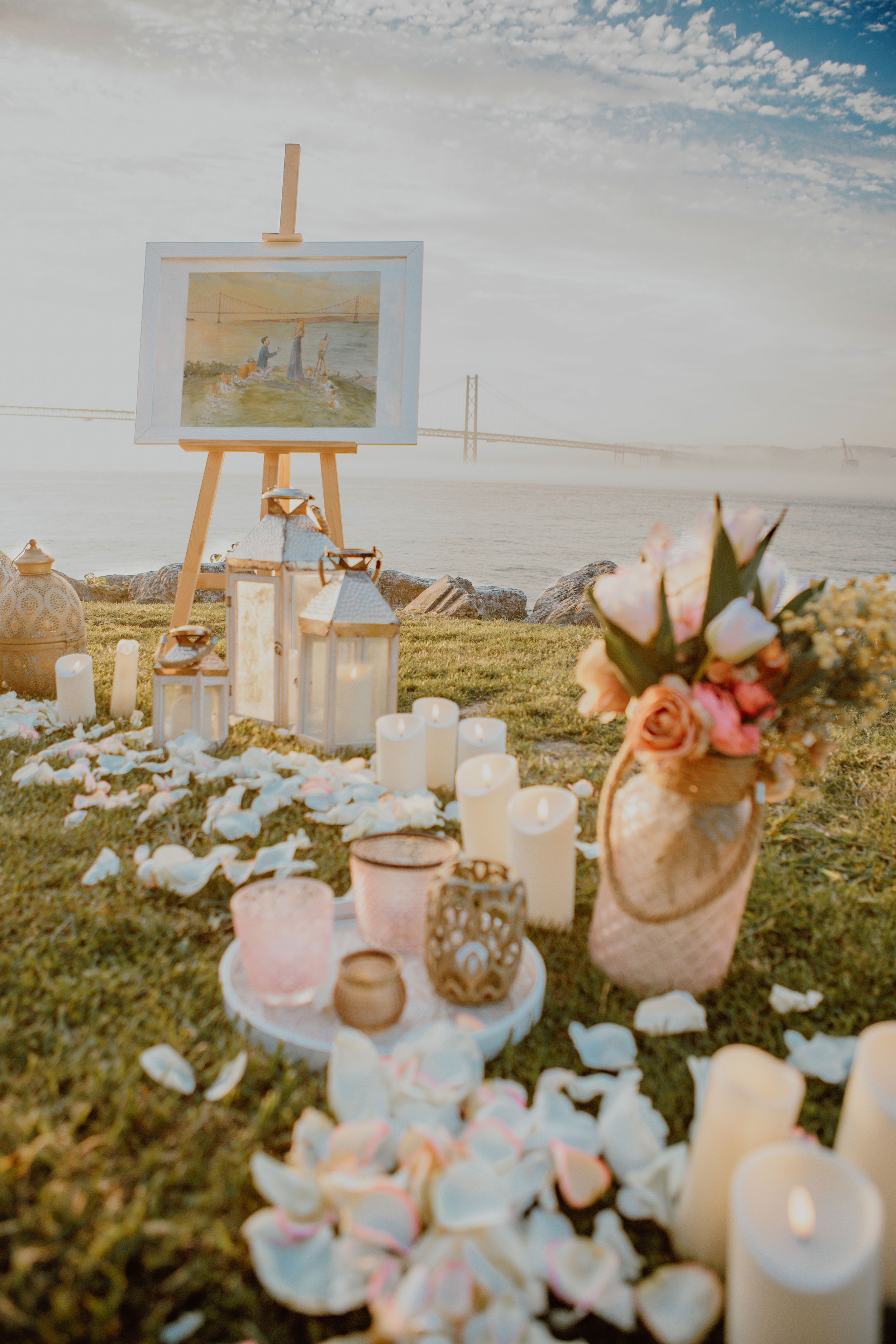 Romantic sunset proposal setup with candles, flowers, and an easel arranged on grassy coastal cliff overlooking the ocean.