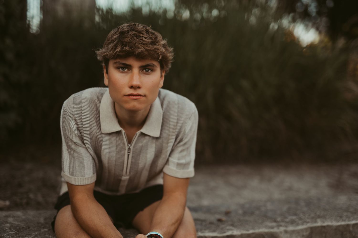 Young man with curly hair wearing a gray zip-up polo shirt, sitting outdoors with a relaxed pose during a senior session at Frame Park.