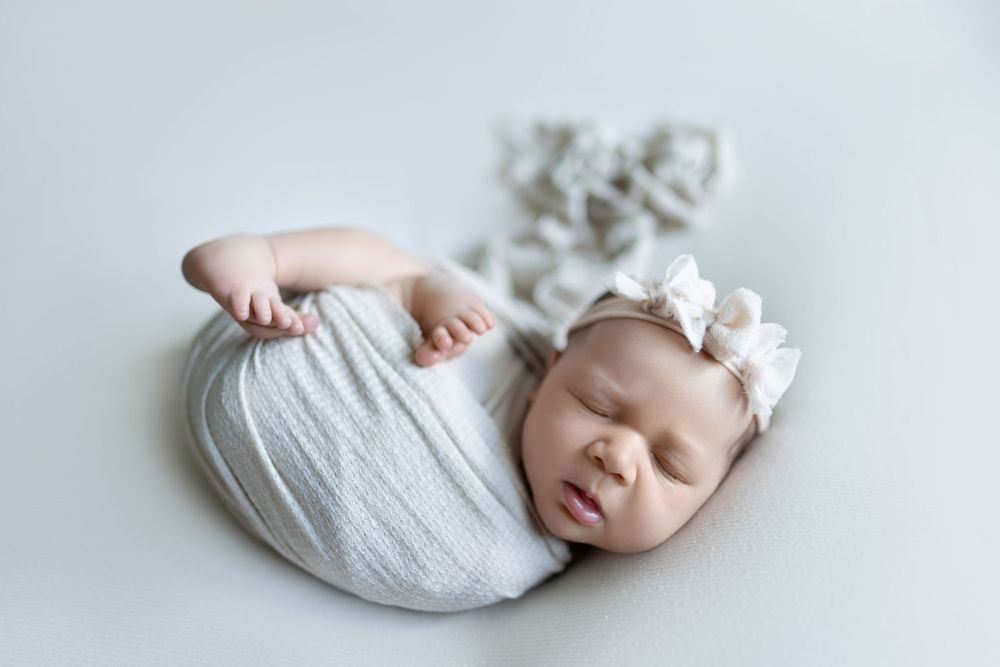 A sleeping newborn baby wrapped in soft white fabric lays peacefully against a light gray background.
