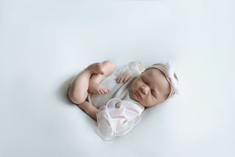 A sleeping newborn baby wearing a white onesie lies peacefully on a soft white blanket.