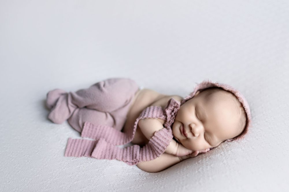 Sleeping newborn baby wearing pink knit hat and wrapped in soft blanket lying on white backdrop.