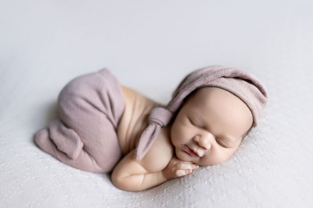 Sleeping newborn baby in soft pink outfit and hat rests peacefully on white backdrop.