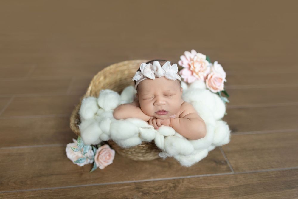A sleeping newborn baby nestled in soft white fabric with delicate pink flowers on a wooden floor.