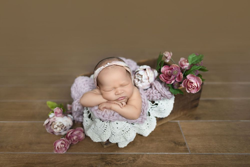 A sleeping newborn baby lies peacefully on a wooden floor surrounded by delicate pink roses and white lace fabric.