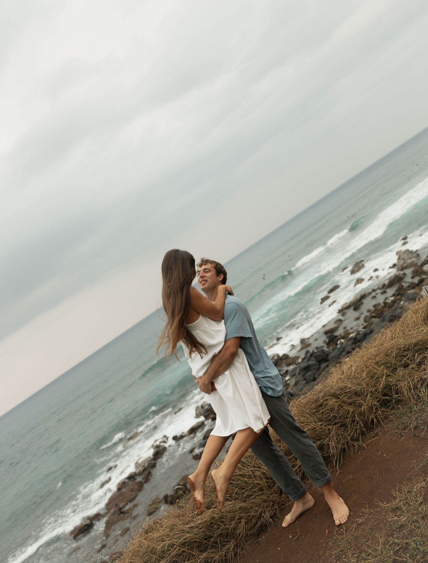 A couple embraces on a sandy beach with ocean waves and cloudy skies in the background.