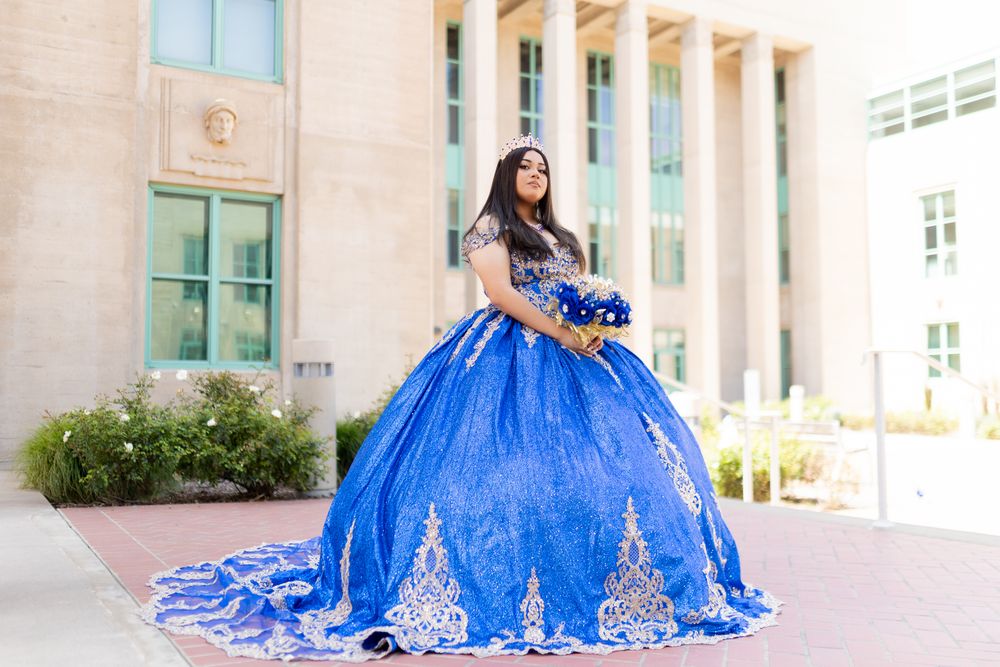 A person in a stunning royal blue and silver quinceañera ball gown poses elegantly in front of a building.