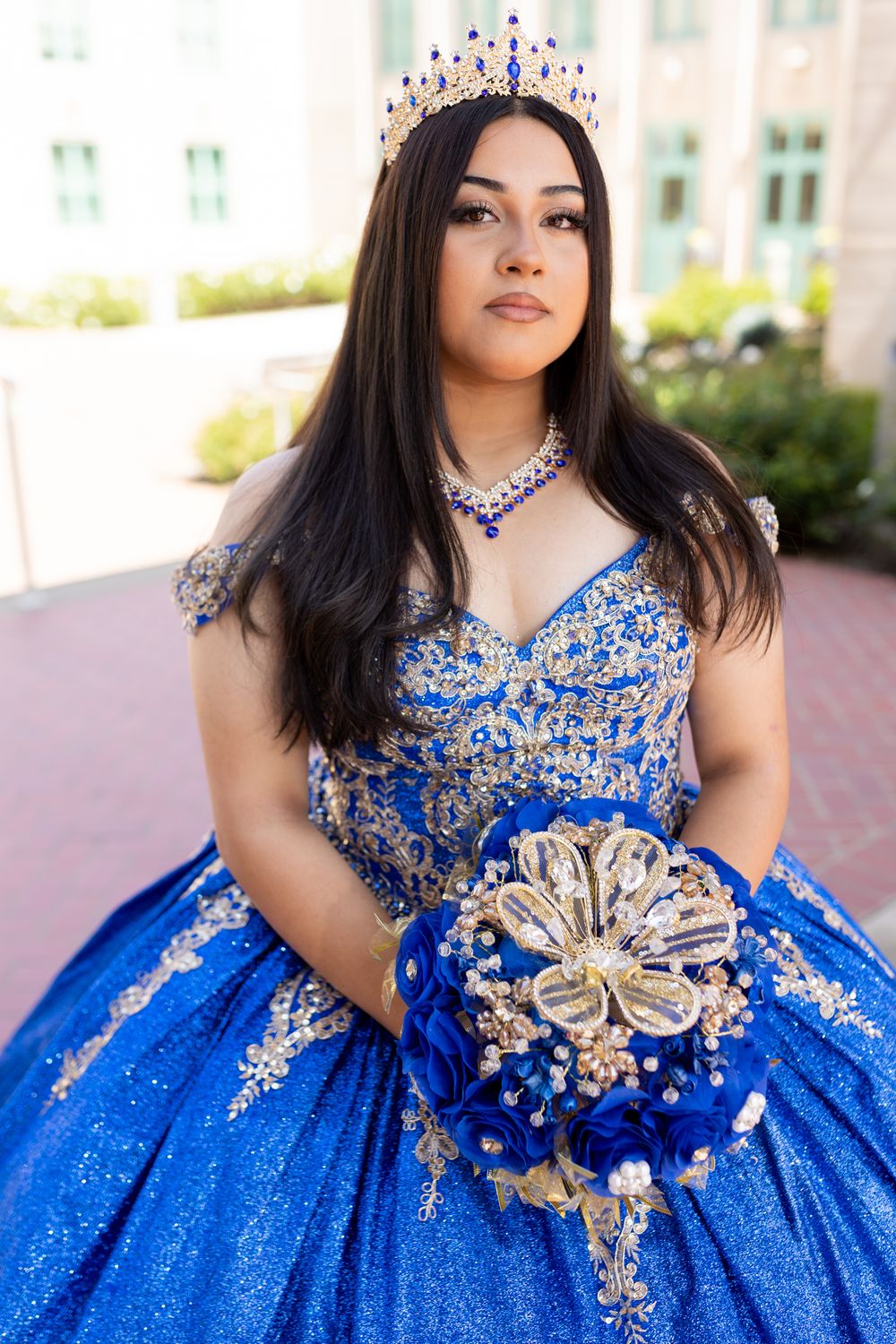 A person in a royal blue quinceañera dress with crystal embellishments and a crown holds a gold and blue bouquet.