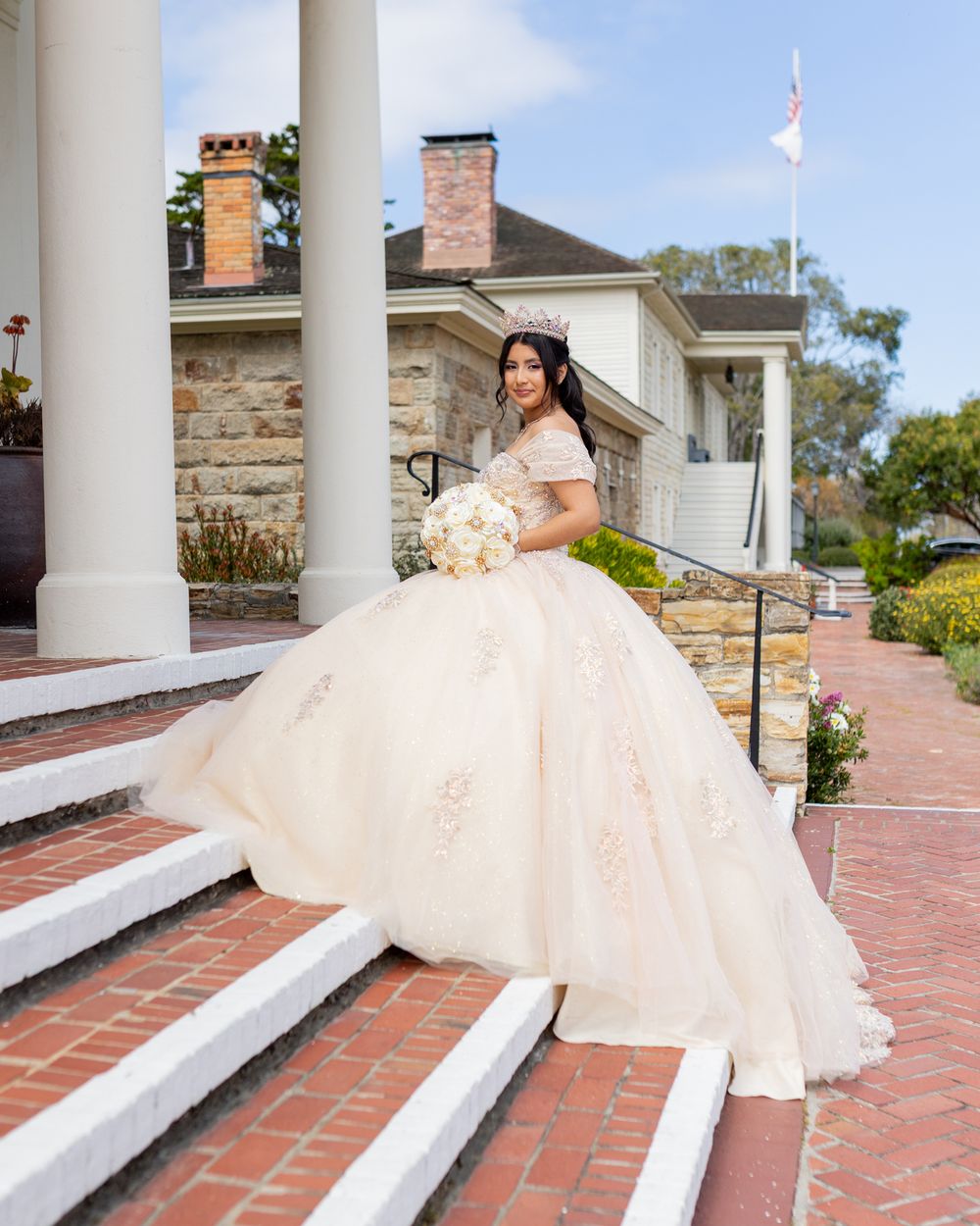 A bride in a cream ball gown sits gracefully on brick steps with white columns and holds a bouquet at an outdoor venue.