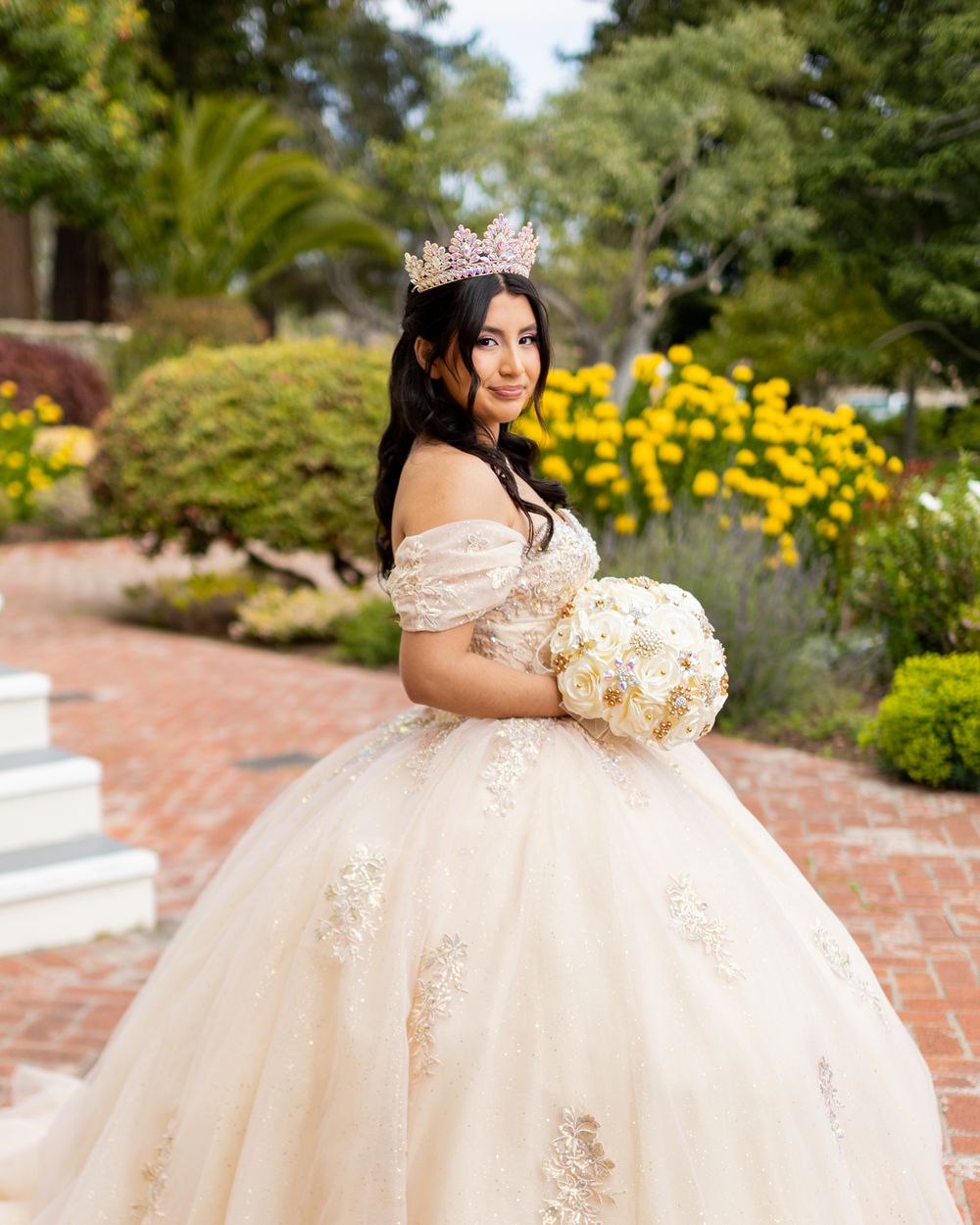 A bride in a sparkly off-shoulder ballgown and tiara poses with a white bouquet in a garden with yellow flowers.