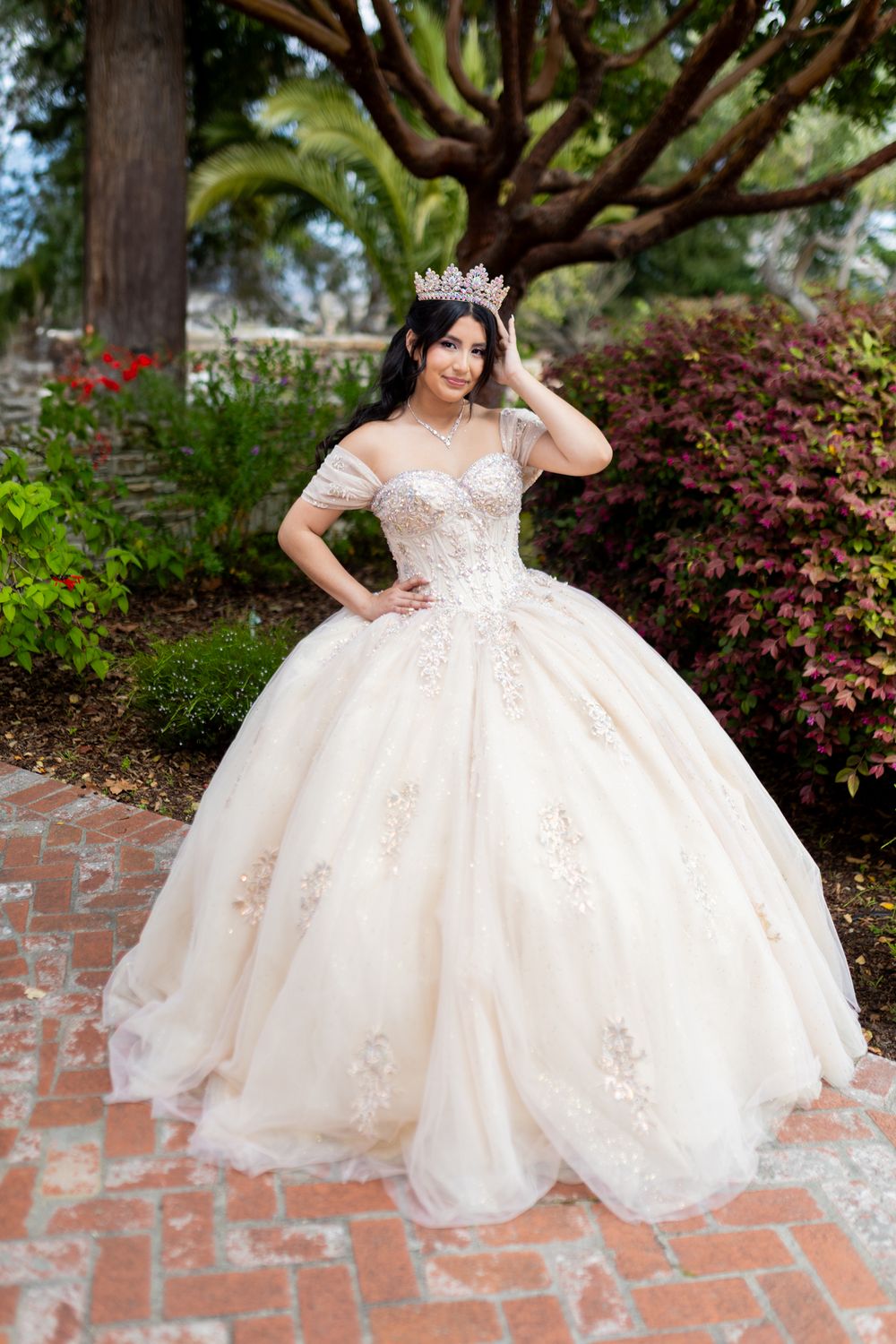 An elegant off-shoulder ballgown with delicate lace appliques stands out against a lush garden backdrop with brick pavers.