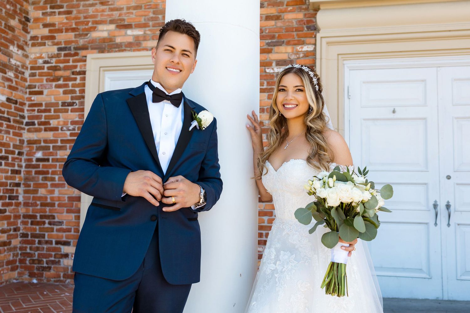 A bride and groom pose together against a brick wall and white column, with the bride holding a white floral bouquet.