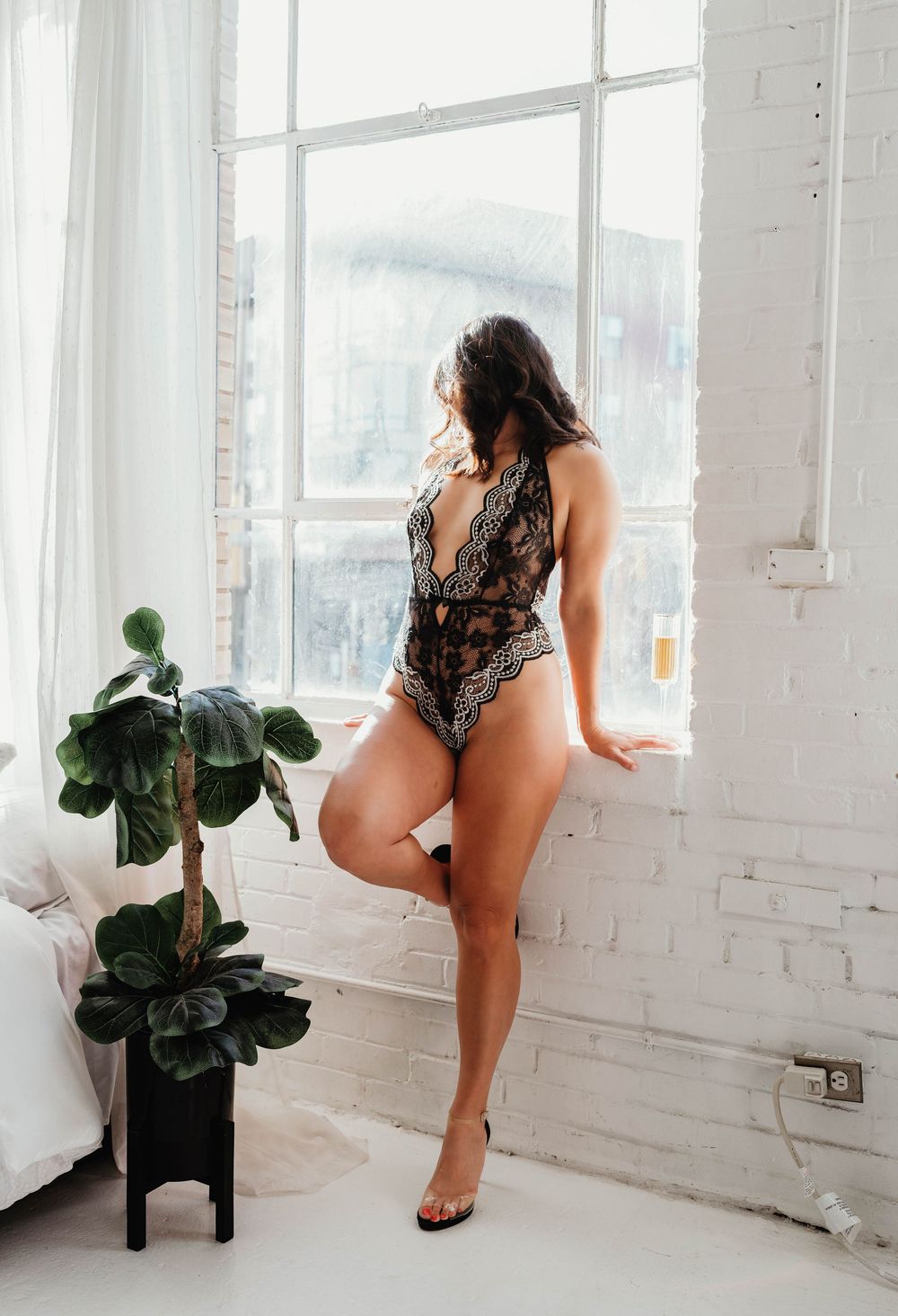Model in black lace bodysuit poses by bright window with decorative houseplants in minimalist white room.