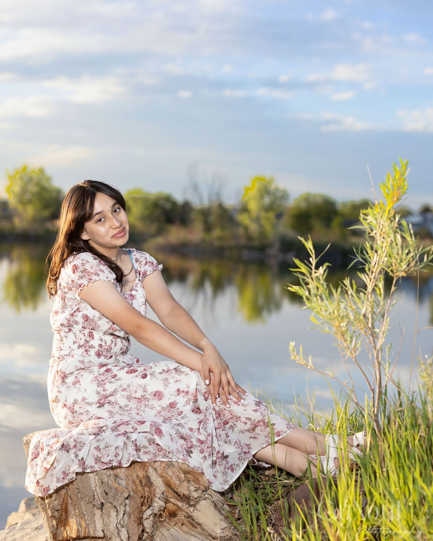 Person in floral dress sits peacefully on tree stump by tranquil lake with tall grasses and autumn trees in background.