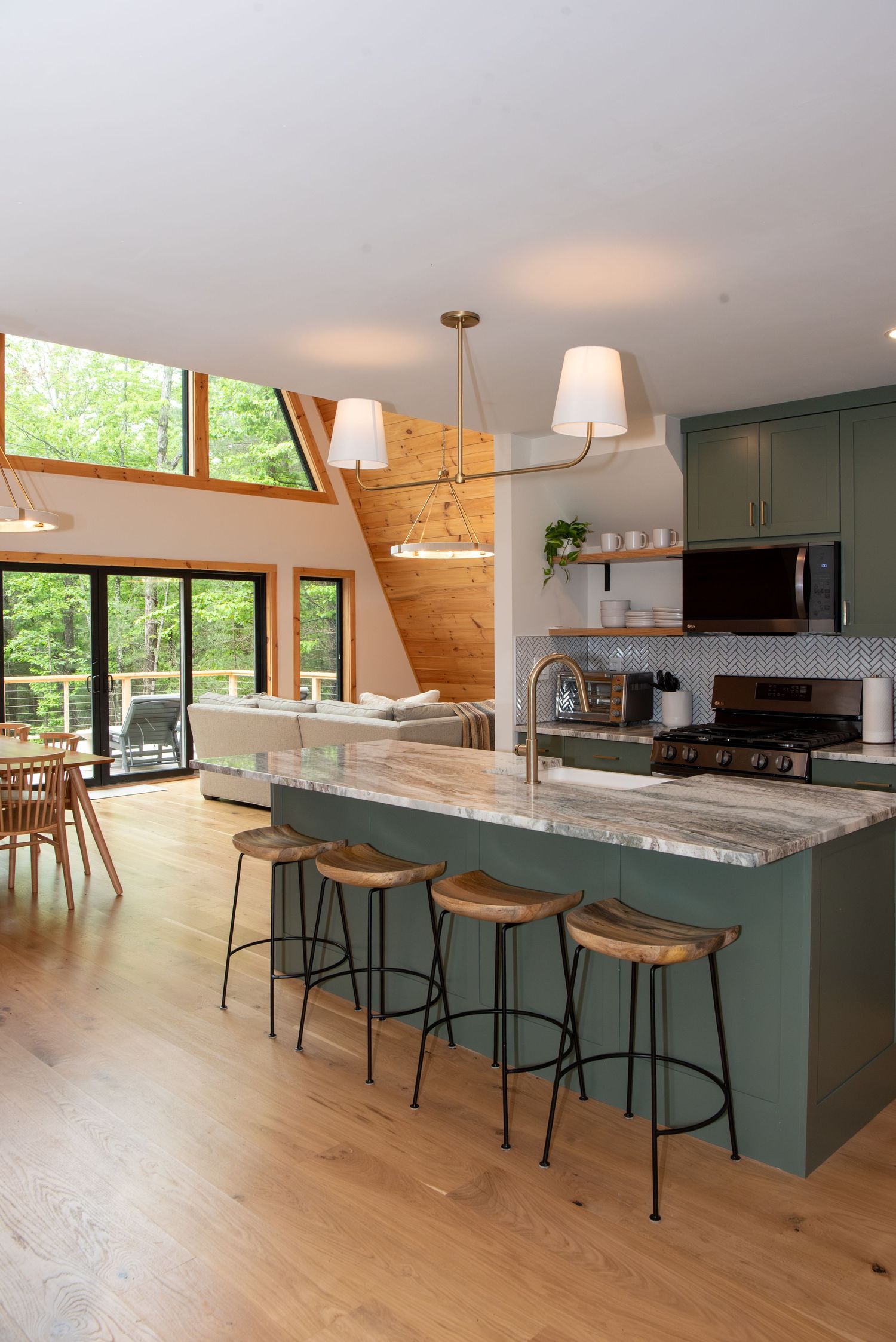 Modern kitchen with sage green cabinets, wood bar stools, and light hardwood floors showcases a spacious open layout.