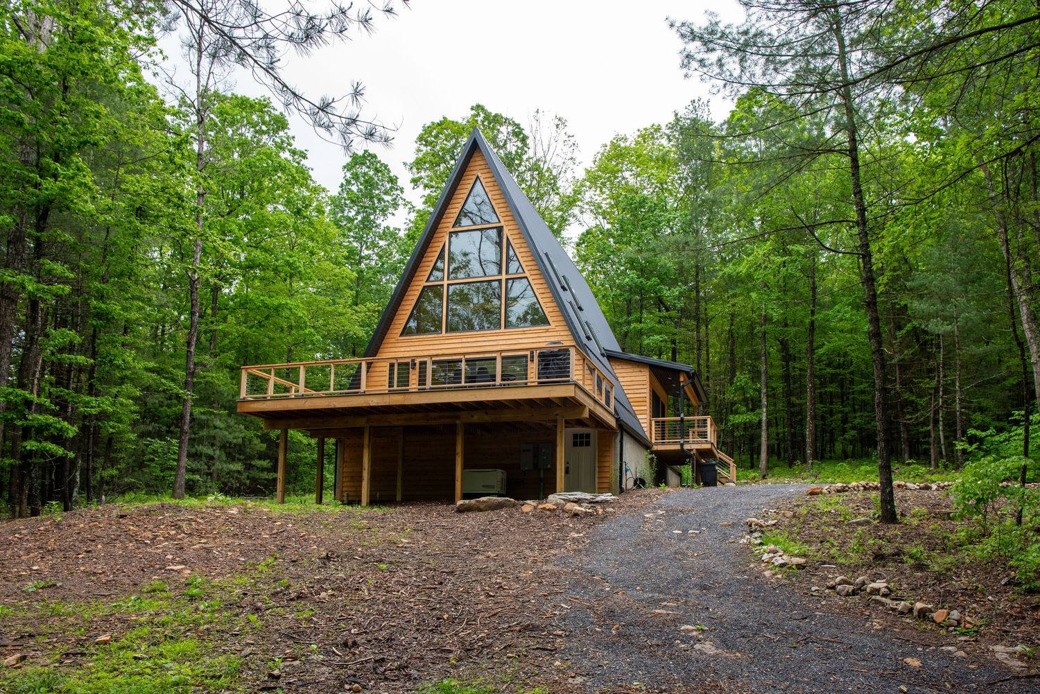 Modern A-frame cabin with large deck nestled in dense forest with gravel driveway approach.