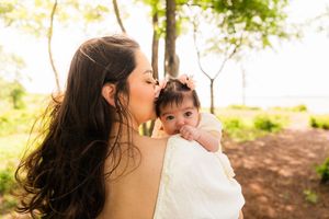 A mother tenderly kisses her baby girl outdoors in a sunlit park surrounded by green trees.