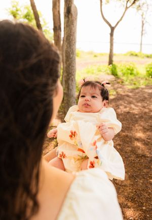 A mother holds her baby girl in a floral dress outdoors among trees in warm golden sunlight.