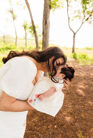 A woman in a white dress tenderly kisses a baby outdoors among trees in warm sunlight.