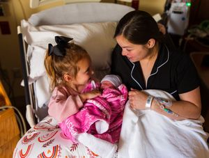 Mother in hospital bed smiles at toddler girl meeting newborn baby sibling wrapped in pink blanket.