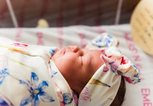 Newborn baby swaddled in a floral blanket with a bow headband sleeping peacefully in a hospital bassinet.