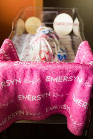 Pink personalized baby blanket with EMERSYN Rose text in a hospital bassinet with newborn care items.