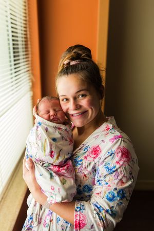 Smiling new mother in floral robe holding her swaddled newborn baby near a window.