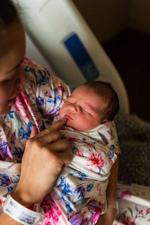 Mother in floral robe cradling swaddled newborn baby in hospital room after birth.
