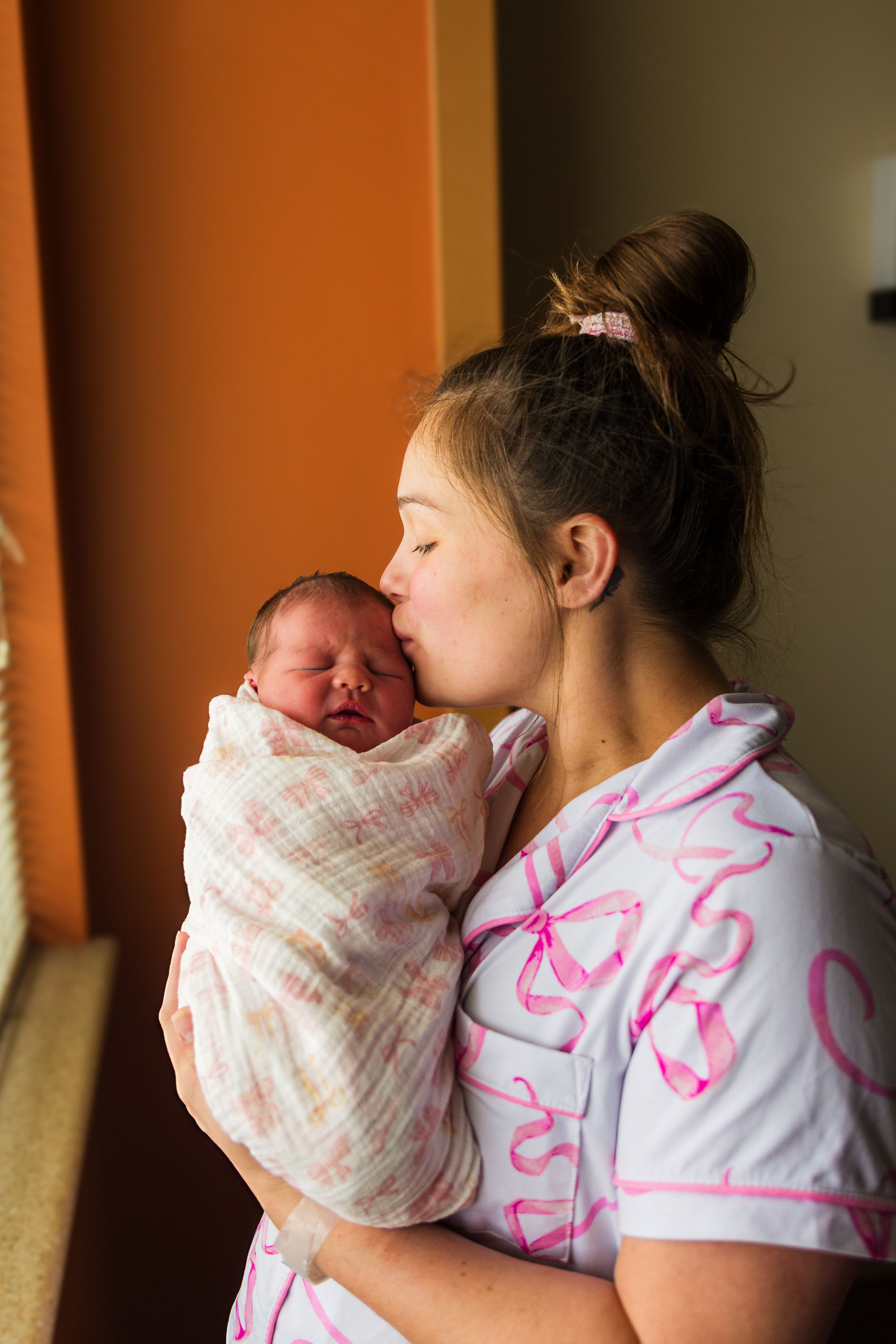 Young mother in pink bow pajamas tenderly kissing her swaddled newborn baby near a window.